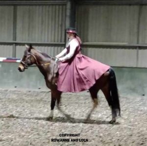 A woman on a dark brown horse, wearing a modern riding dress inspired by Anglo Saxon riding horse.