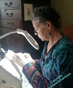 Me, a white woman with greying hair, working at an embroidery frame held under a magnifer and working light.